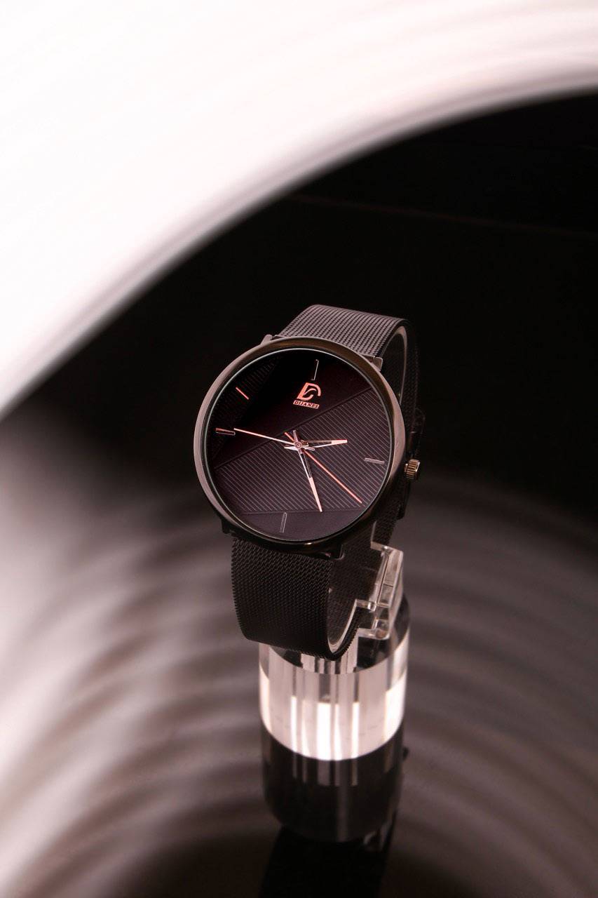 Montre Quartz Unisexe - Minimaliste Décontracté avec cadran noir épuré et bracelet en maille noire, posée sur un support transparent élégant. Mouvement à quartz précis, idéale pour un style moderne et discret.