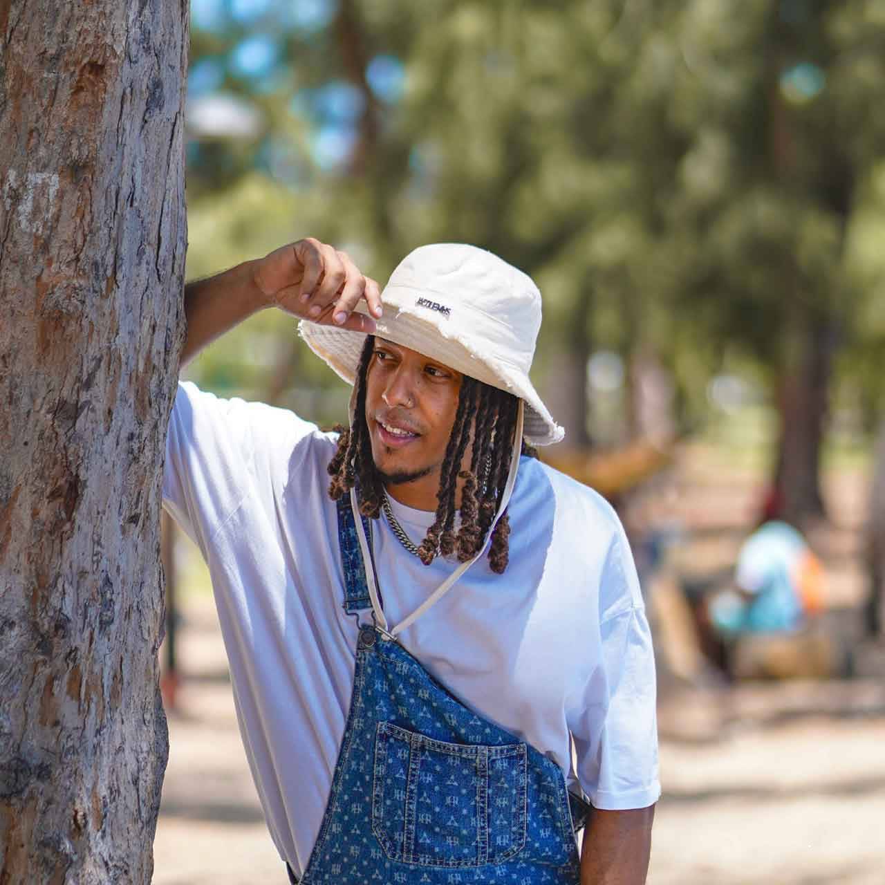 Bob Jacquemus effiloché en coton blanc, ajustable avec logo argenté, porté par un homme en extérieur pour un style tendance et décontracté.